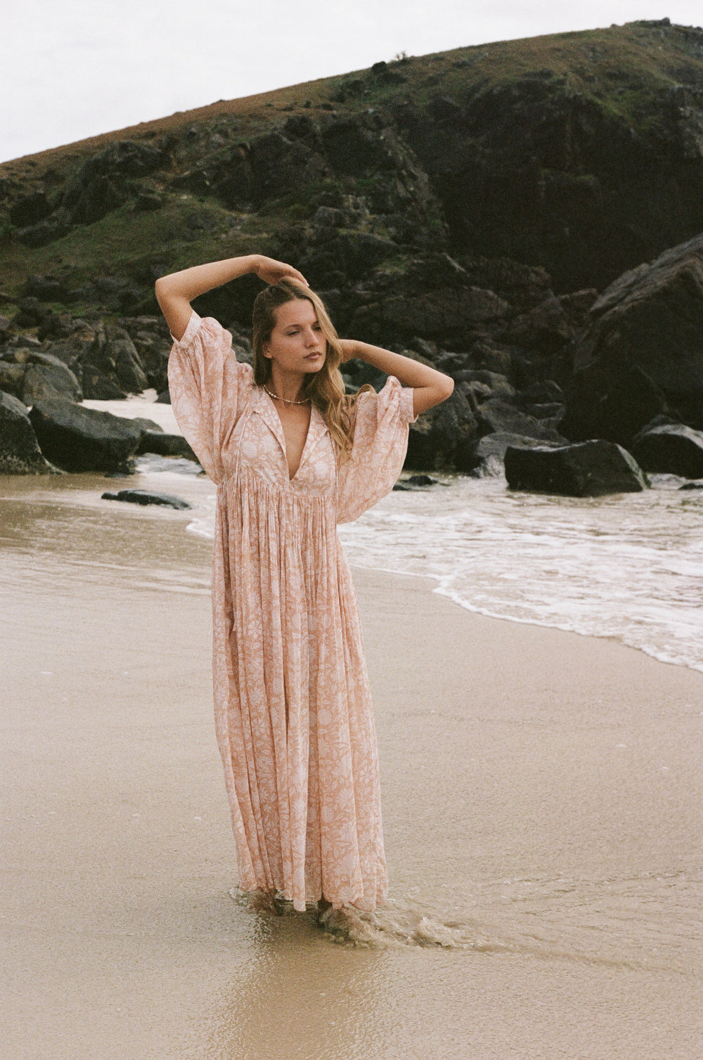 Daughters of India Kyra Maxi Dress in Apricot Blush — standing in shallows arms raised, rocky headland backdrop