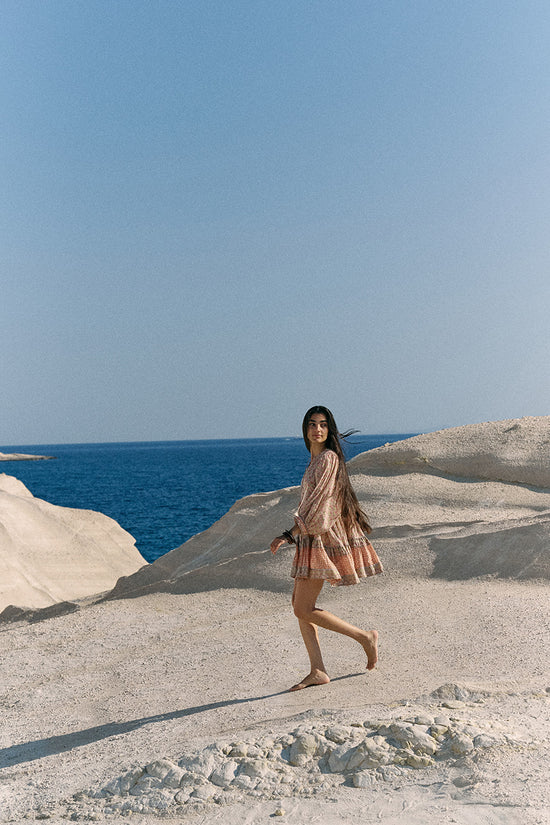 Woman walking across white coastal rock landscape with blue ocean wearing the Daughters of India Bhoomi Mini Dress in Sunset, full-length view