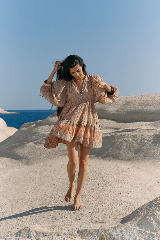 Woman on coastal rock with ocean and blue sky wearing the Daughters of India Bhoomi Mini Dress in Sunset, showing the tiered boho silhouette with flowing hem