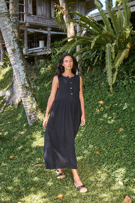 Nadi Dress Black — full length front view in tropical garden, black double gauze cotton midi dress with wooden button front and gathered waist, palm trees and stilt house | Daughters of India