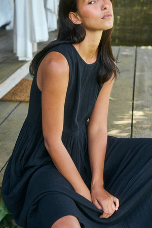 Nadi Dress Black — seated in tropical garden, black double gauze cotton midi dress, relaxed lifestyle pose with greenery | Daughters of India