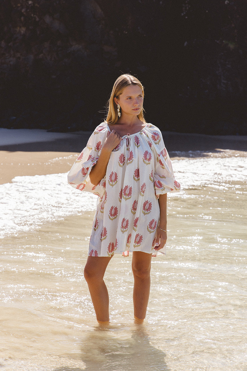 Ria Mini Dress Strawberry Cream — ocean wading lifestyle shot, model standing in shallow waves wearing strawberry cream floral hand block print cotton mini dress, summer beach styling, artisan Jaipur India | Daughters of India