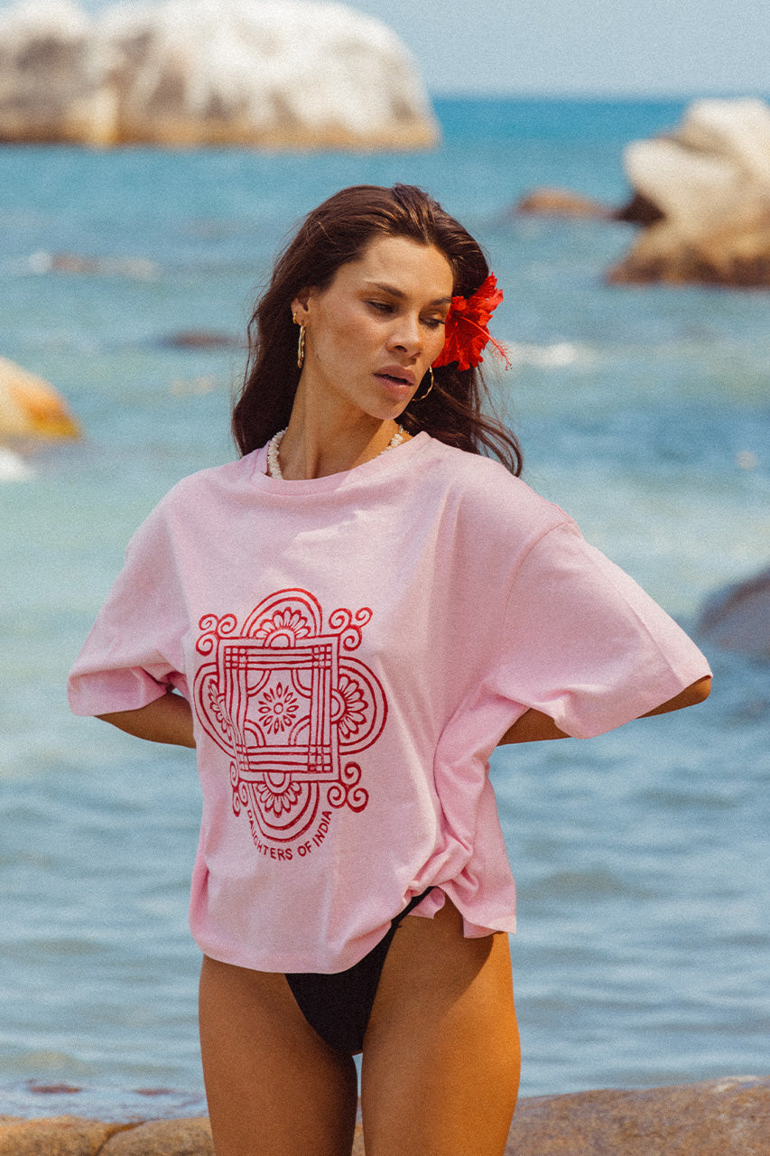 Shanti Tee Pink — close-up upper body portrait at beach, pink oversized cotton tee with red mandala logo, hibiscus in hair, ocean rocks | Daughters of India